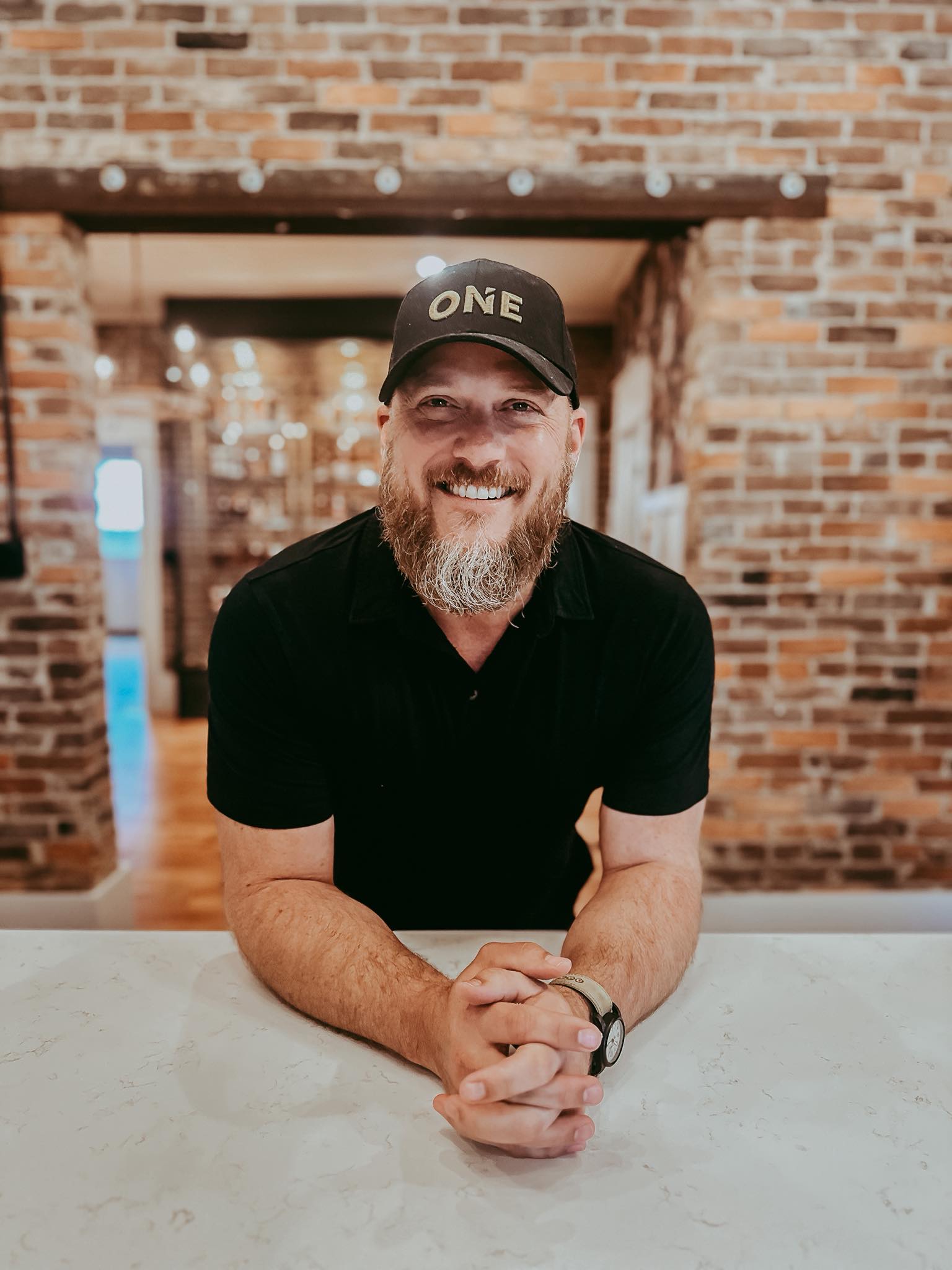 Smiling man with a gray beard and black cap reading 'ONE', leaning on a marble counter in a brick-walled space.