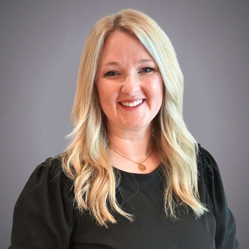 Access Headshots 1 - Access Title & Escrow Smiling woman with long blonde hair wearing a black blouse against a gray background, head-and-shoulders portrait.