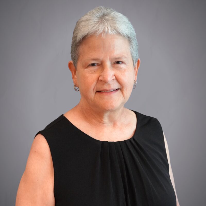 Access Headshots 2 - Access Title & Escrow Professional portrait of an older woman with short gray hair, wearing a black sleeveless top, smiling against a gray backdrop.