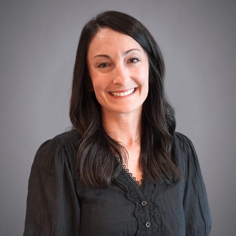 Access Headshots - Access Title & Escrow Smiling woman with dark hair wearing a black blouse, posed against a neutral gray backdrop.